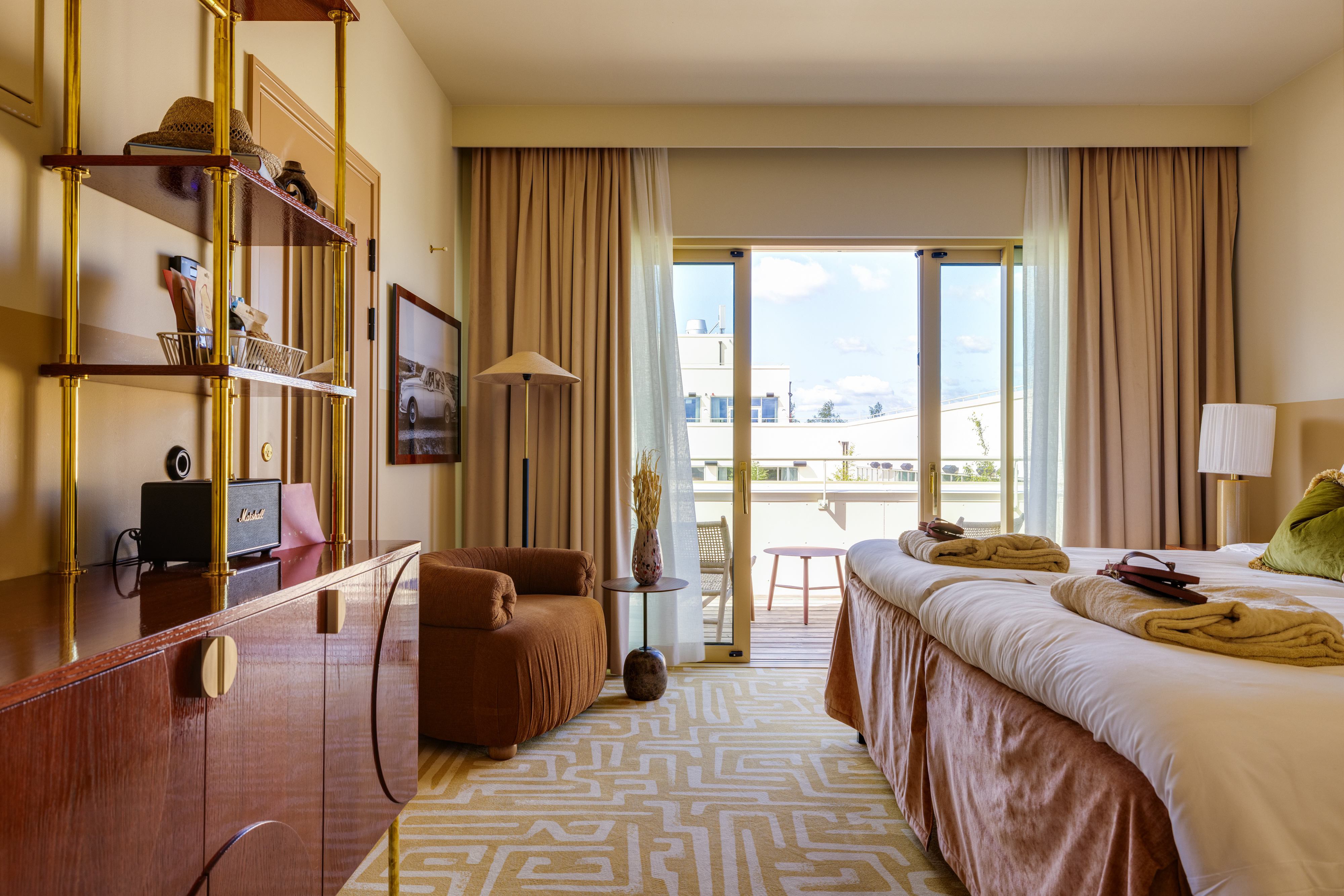 Hotellrum med två bäddade sängar, fåtölj, mässingshylla och balkongdörr med utsikt