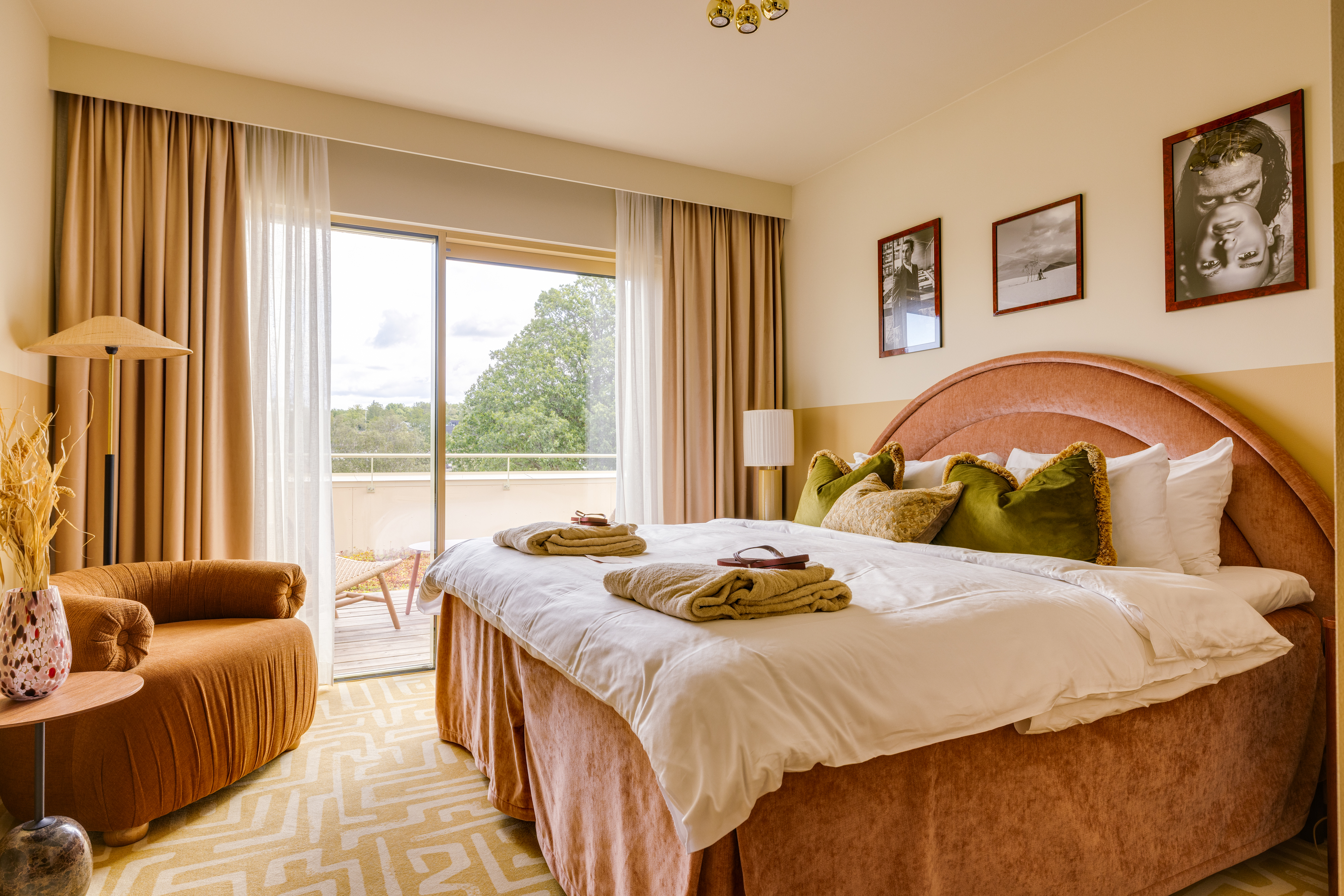 Hotellrum med dubbelsäng, rosa sänggavel, fåtölj och balkongdörr mot grönska