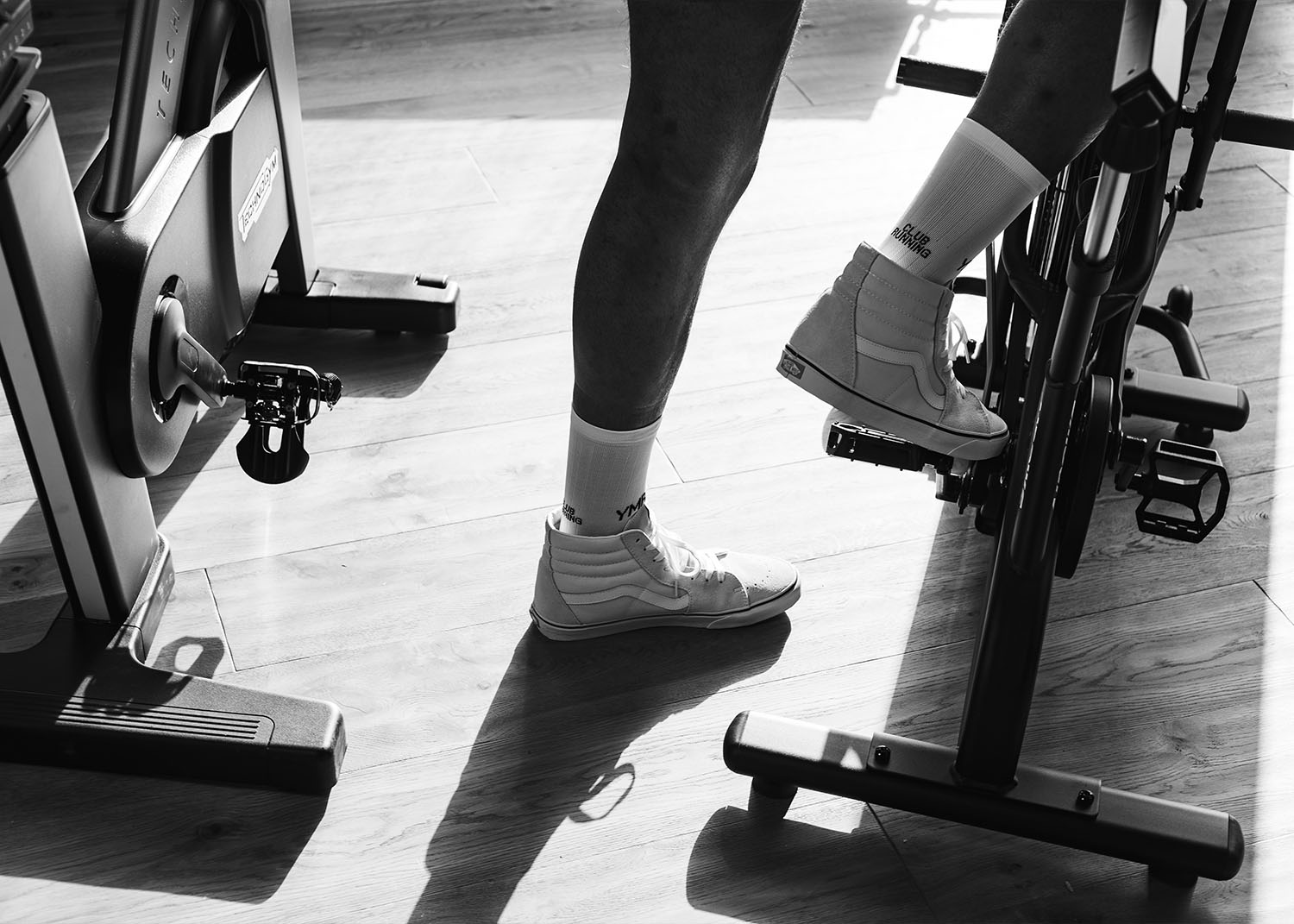 Ben i sneakers på en spinningcykel i en solig träningssal, svartvitt