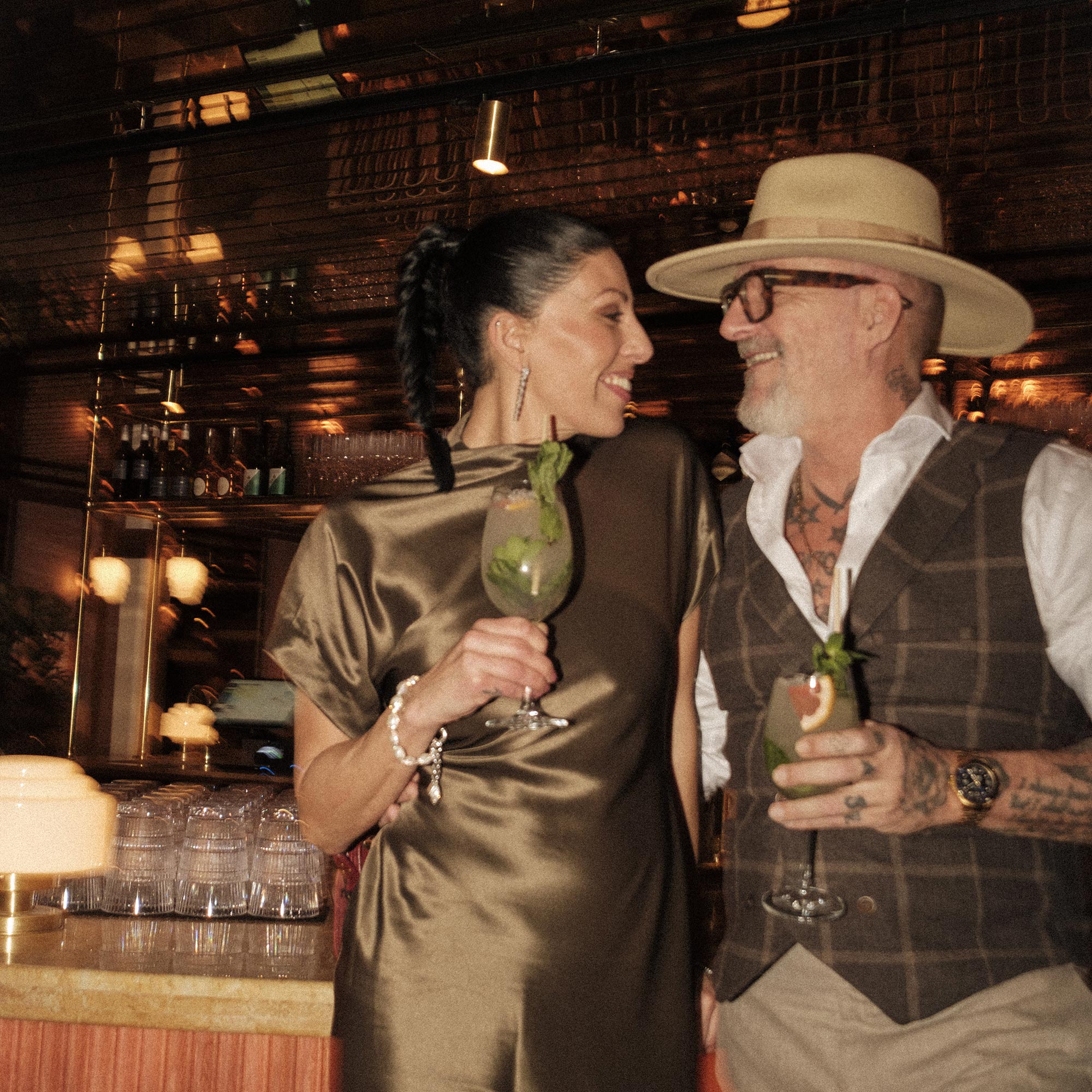 Par i baren med cocktails, mannen i hatt och väst, flaskhyllor i bakgrunden